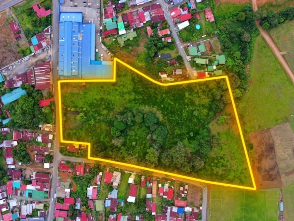 Aerial view of a large grassy lot outlined in bright yellow, bordered by a residential area and open fields behind and to the sides for context.