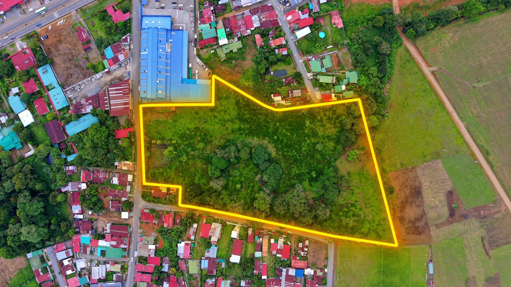 Aerial view of a large grassy lot outlined in bright yellow, bordered by a residential area and open fields behind and to the sides for context.