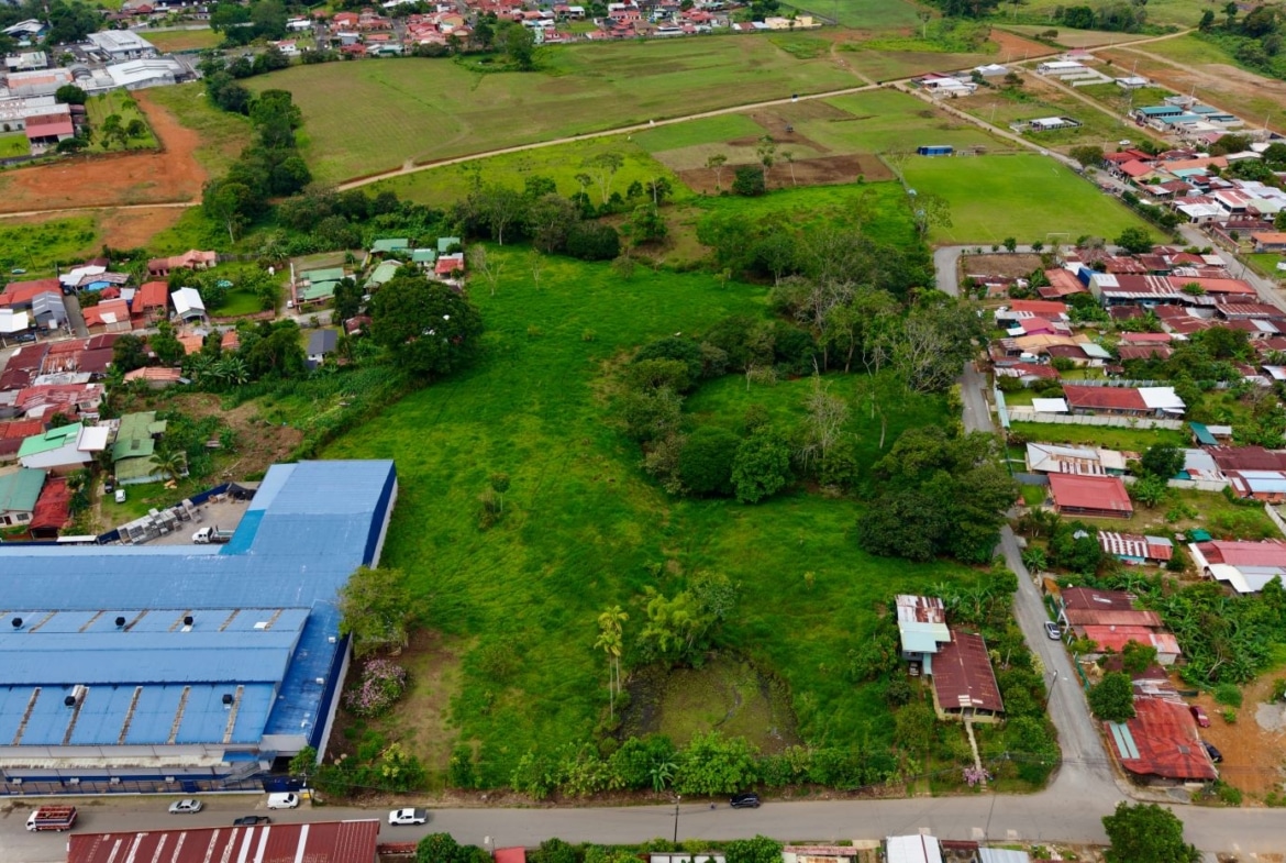 Aerial view of a small town showing a large blue-roofed building on the left, green fields in the center, and residential streets with houses on the right.