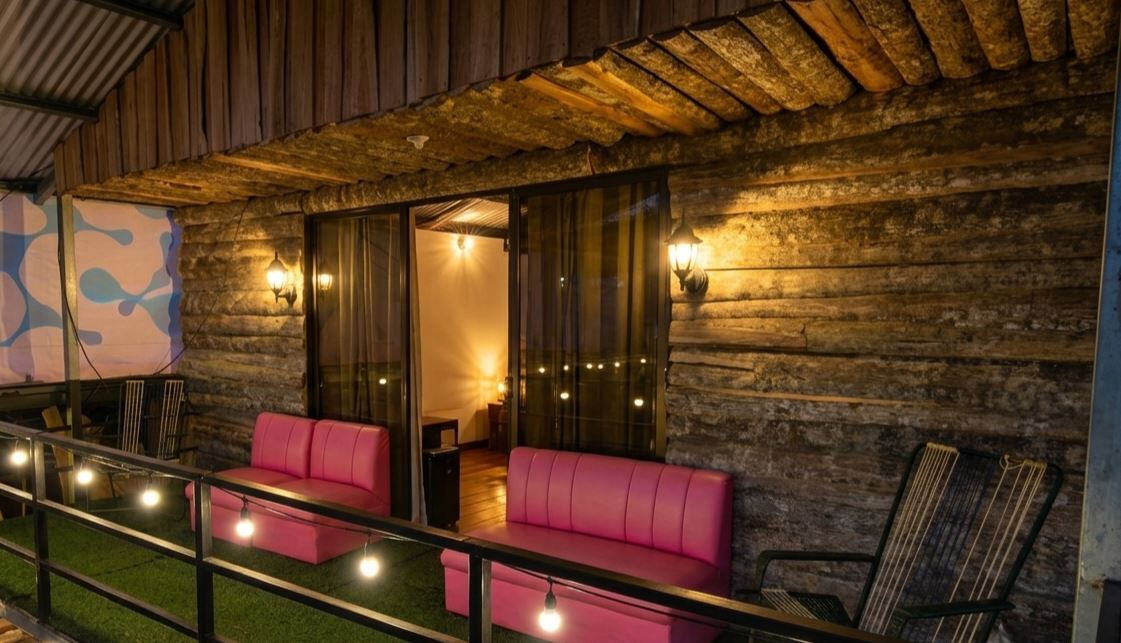 Cozy outdoor patio on a wooden balcony with pink cushioned benches, string lights, and a sliding glass door leading to a warmly lit interior room at night.