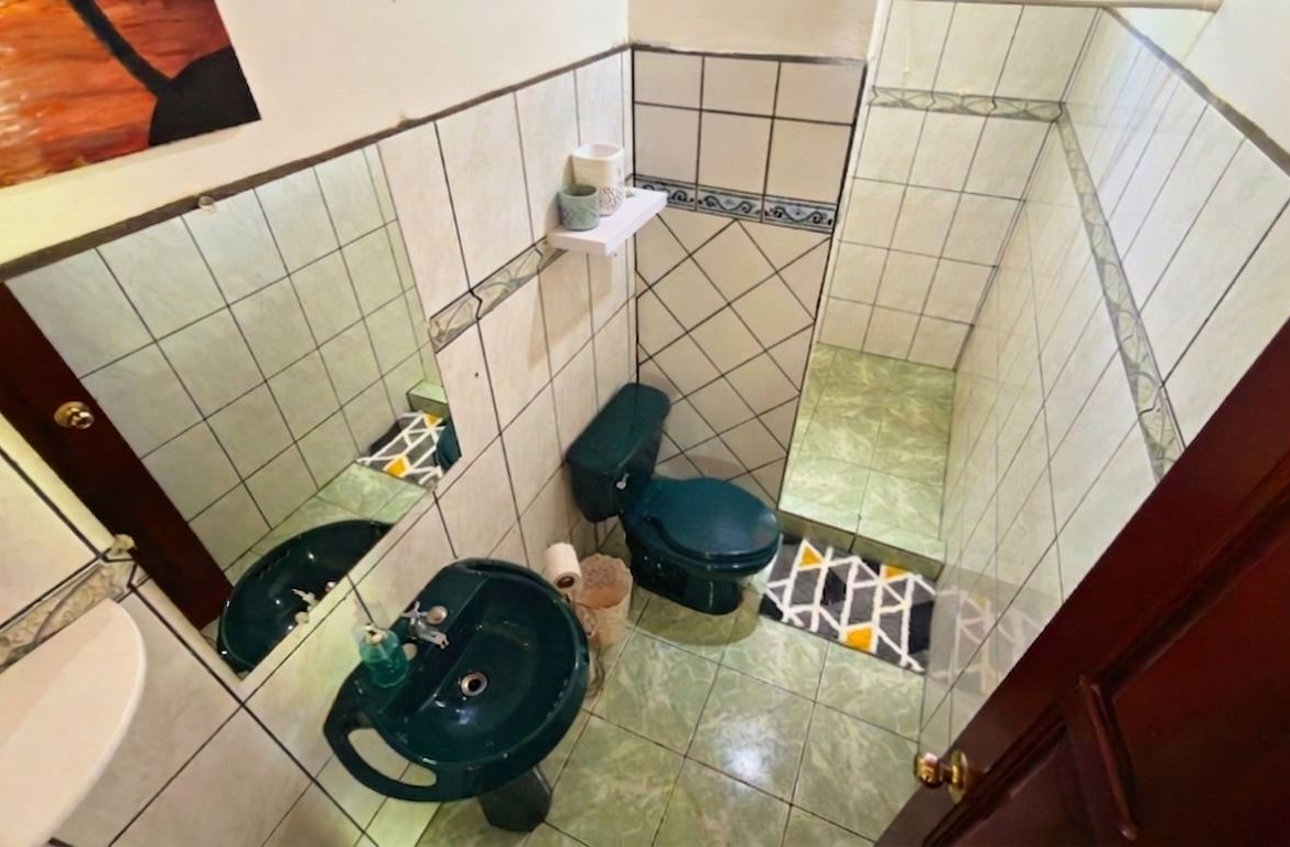 Small teal bathroom with toilet and sink, tiled walls and floor, mirror on the wall, and a shelf holding toilet paper above the toilet.