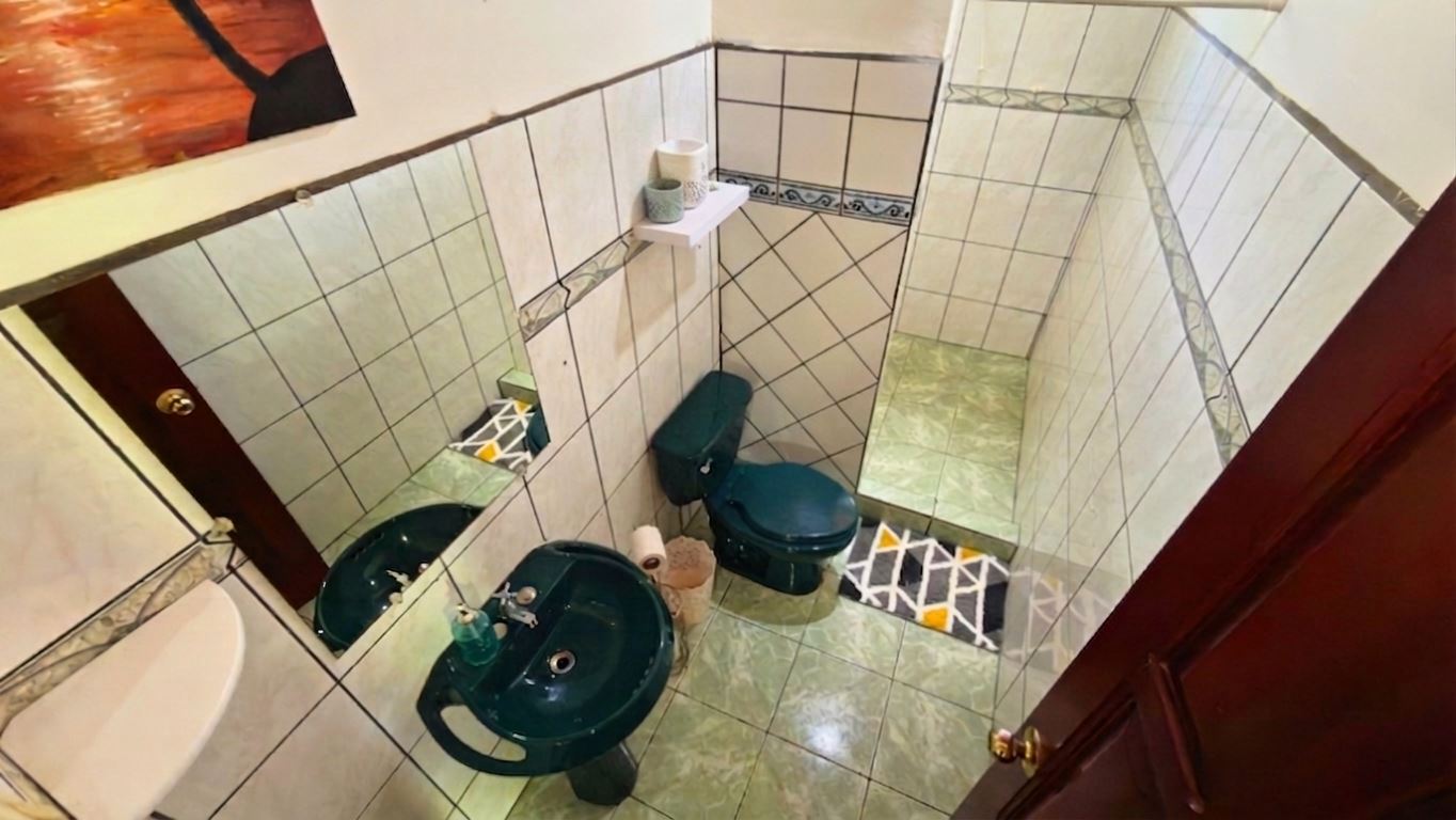 Small teal bathroom with toilet and sink, tiled walls and floor, mirror on the wall, and a shelf holding toilet paper above the toilet.