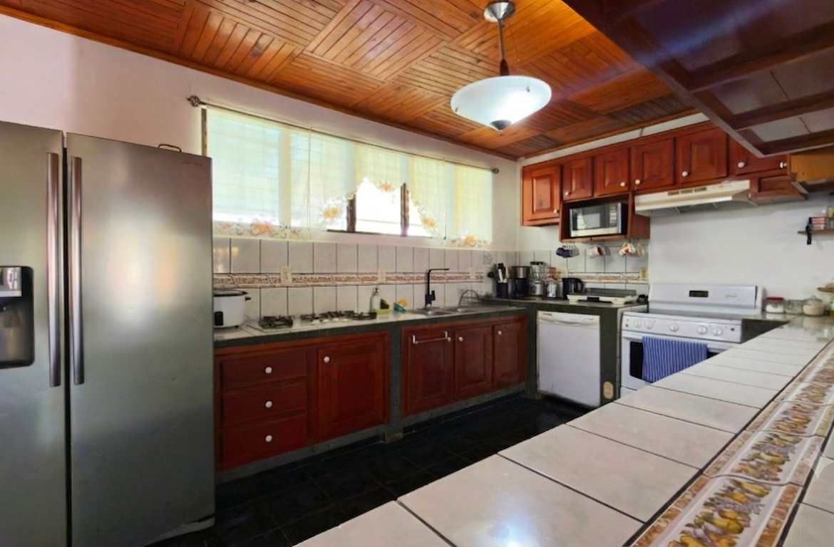 L-shaped kitchen with wooden cabinets, a stainless steel refrigerator on the left, gas stove, and tiled counter in the foreground under a wooden ceiling.