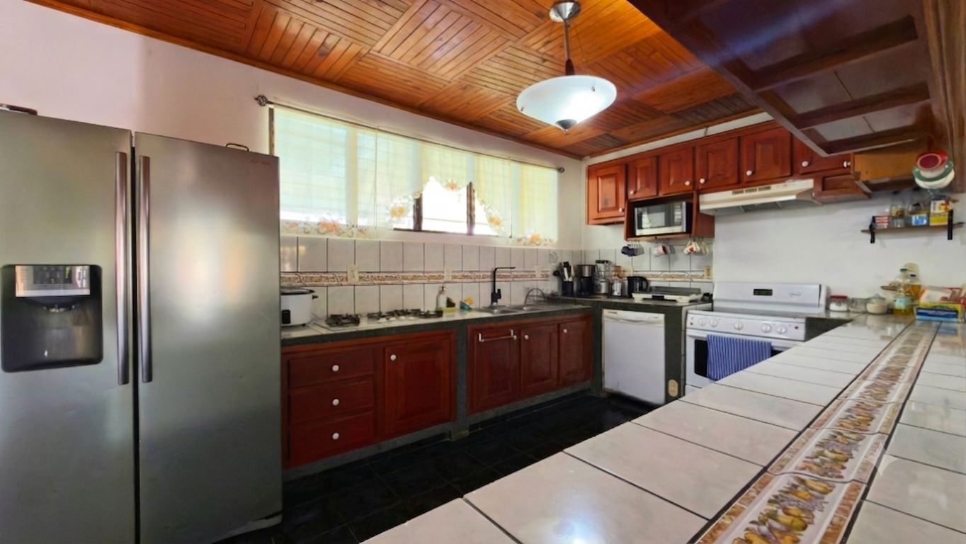 L-shaped kitchen with wooden cabinets, a stainless steel refrigerator on the left, gas stove, and tiled counter in the foreground under a wooden ceiling.