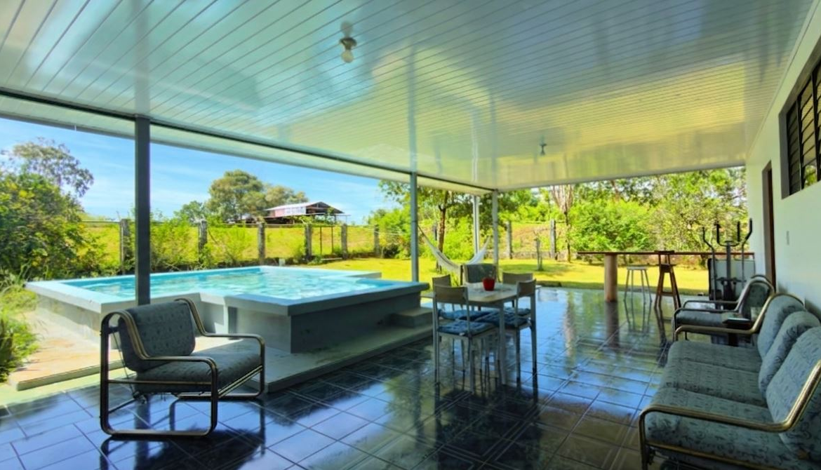 Covered patio over a tiled floor with a raised rectangular pool, outdoor dining table, hammock, and cushioned seating along a fence-backed yard.