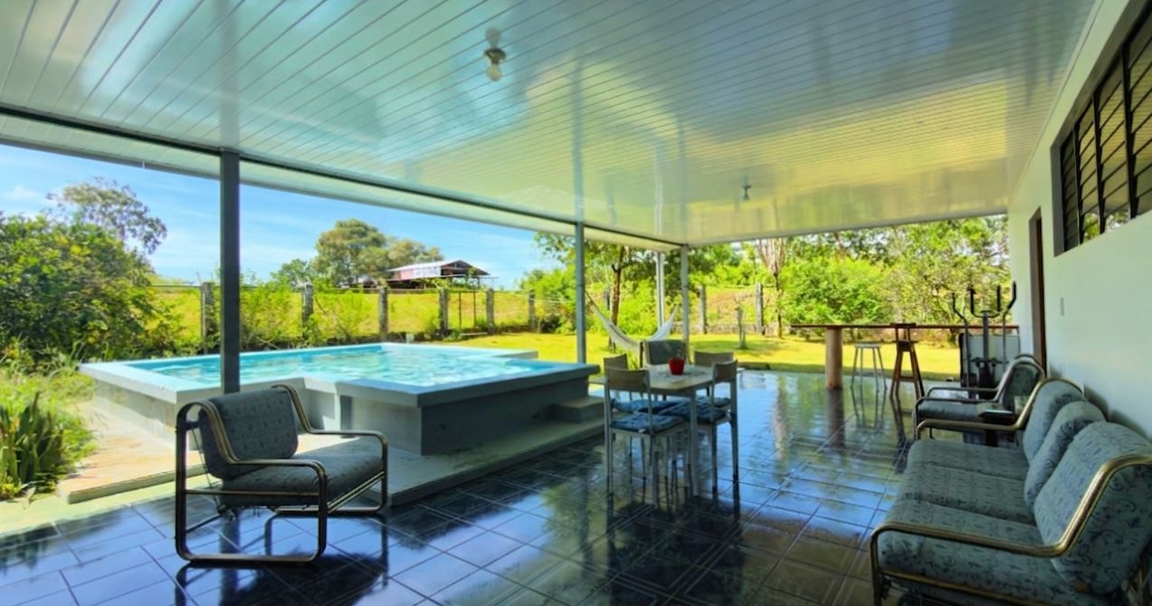 Covered patio over a tiled floor with a raised rectangular pool, outdoor dining table, hammock, and cushioned seating along a fence-backed yard.