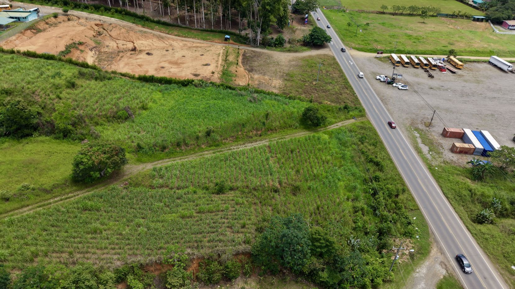 Aerial view of green farmland with planted rows, a diagonally crossing paved road, and a yard of parked school buses, trailers, and trucks on the right.