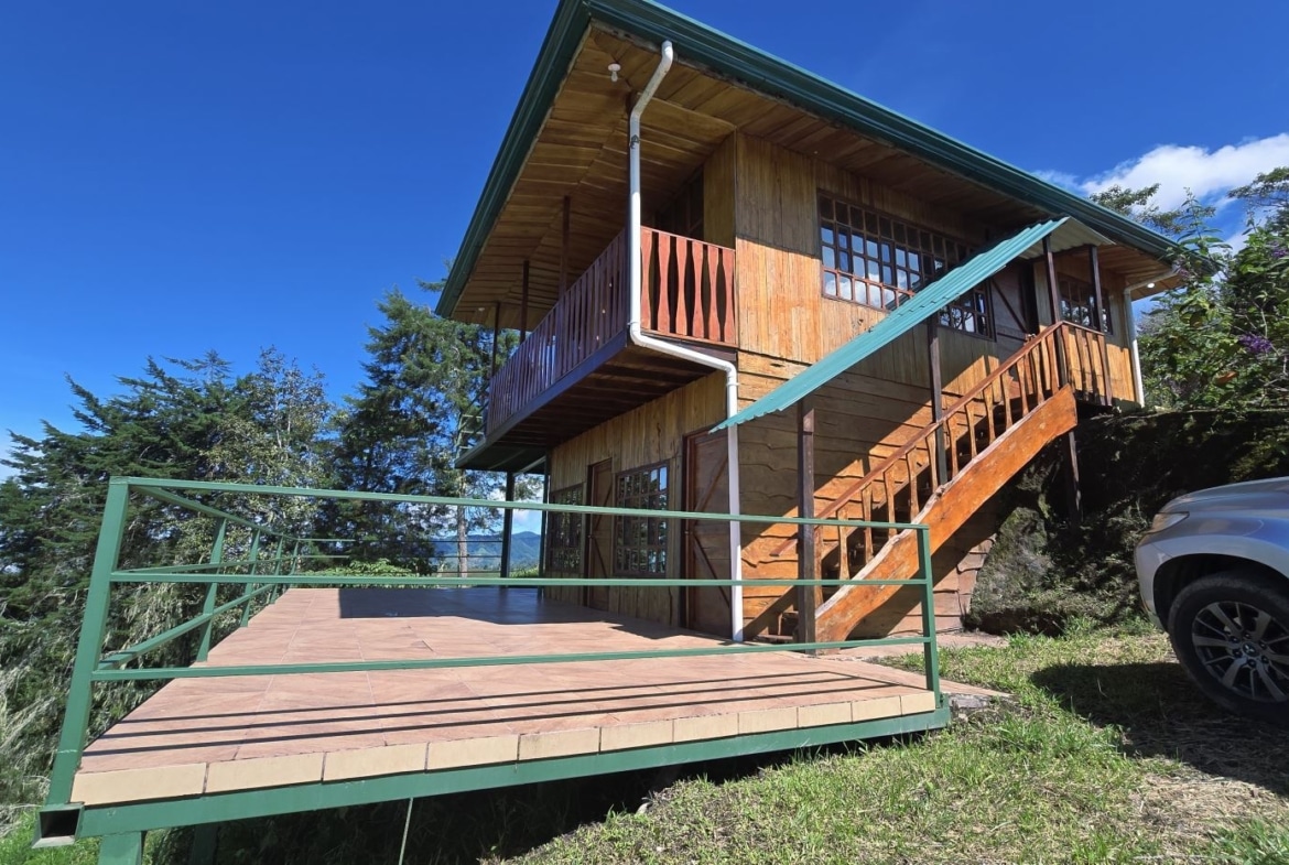 Wooden two-story house with a large green-railed deck on a hillside, stairs to the front, under a blue sky; a silver SUV parked nearby on the right.