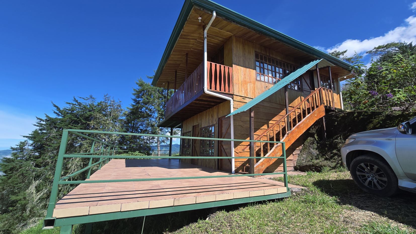 Wooden two-story house with a large green-railed deck on a hillside, stairs to the front, under a blue sky; a silver SUV parked nearby on the right.