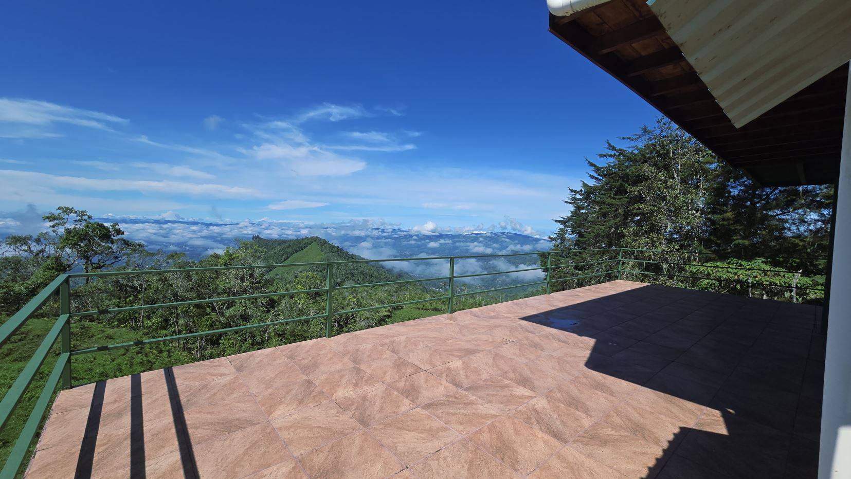Terrace with brown tiles and green railing, overlooking green mountains and a blue sky with clouds.
