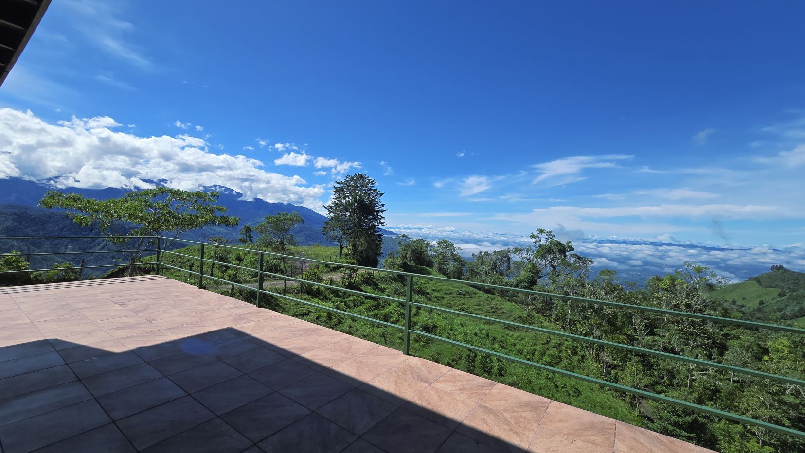 Terrace with a tiled floor and green railing overlooking a valley of trees and mountains under a bright blue sky.