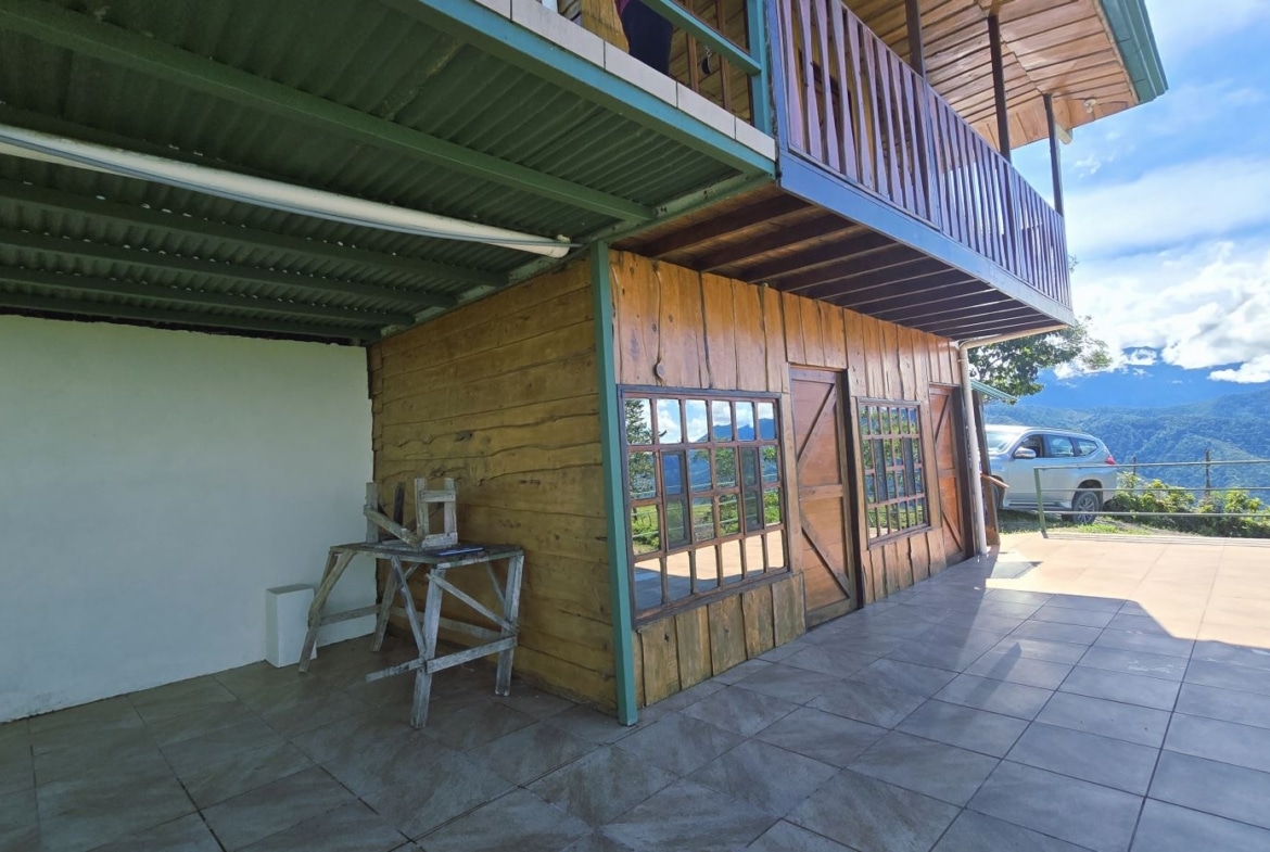 Terraced wooden house with large windows on a tiled patio, a car parked by a railing, overlooking mountains and blue sky.