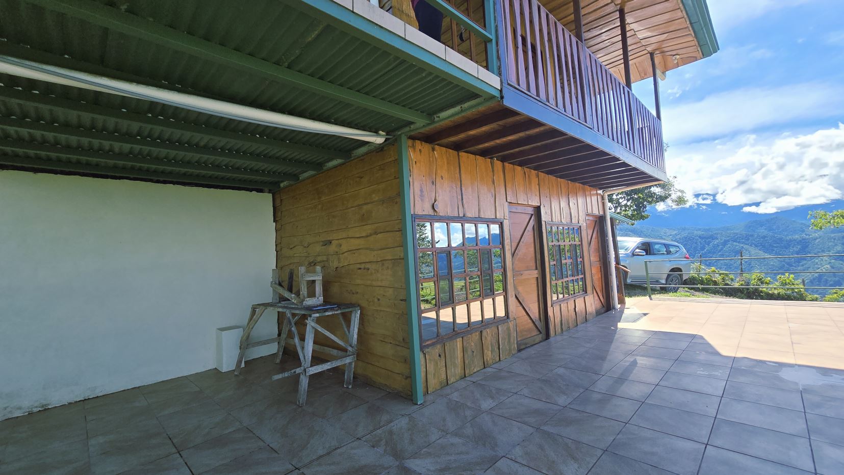 Terraced wooden house with large windows on a tiled patio, a car parked by a railing, overlooking mountains and blue sky.