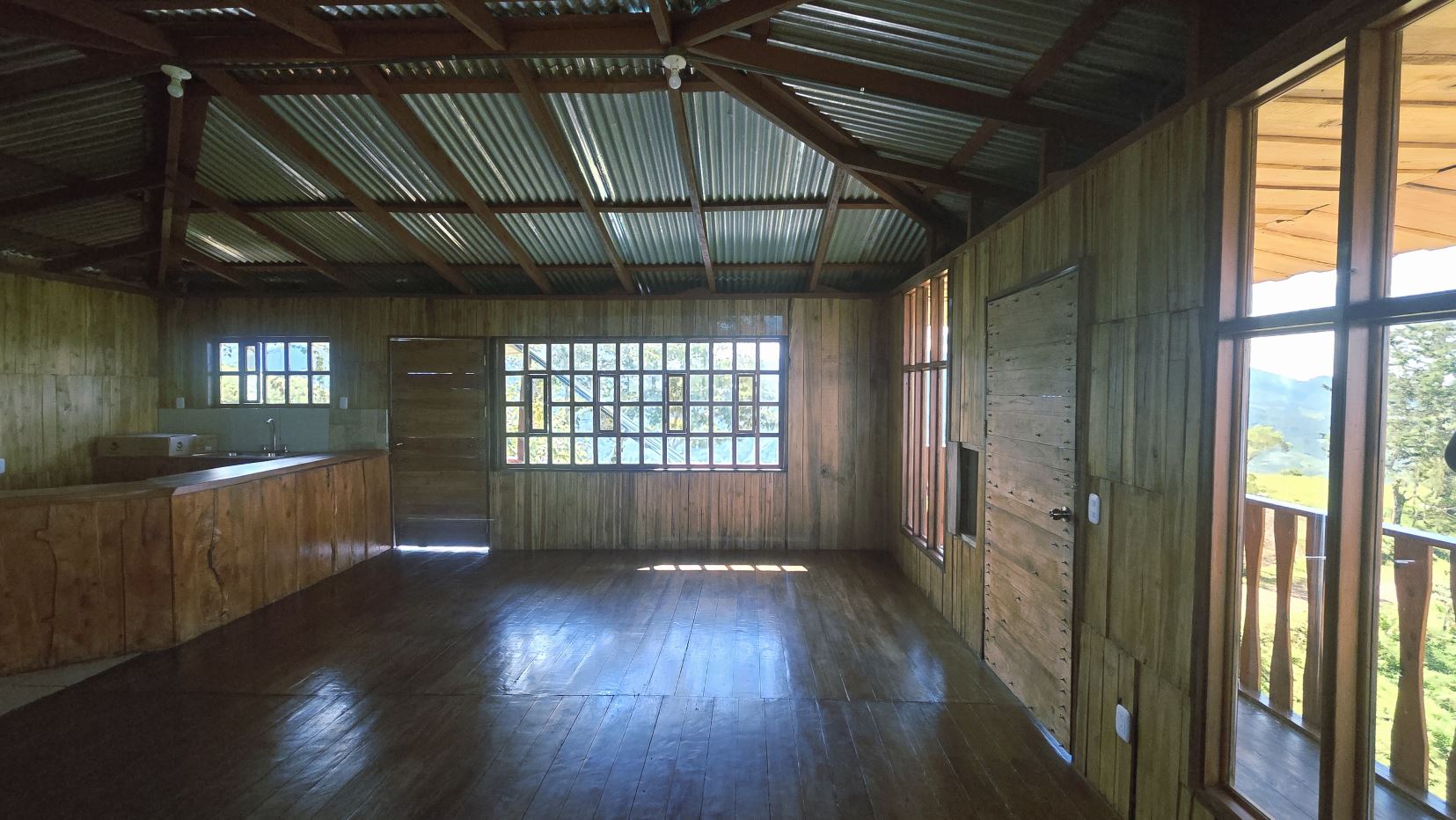Ru stic wooden interior with a curved kitchen counter on the left, wood-paneled walls, and a large window revealing a green landscape outside.