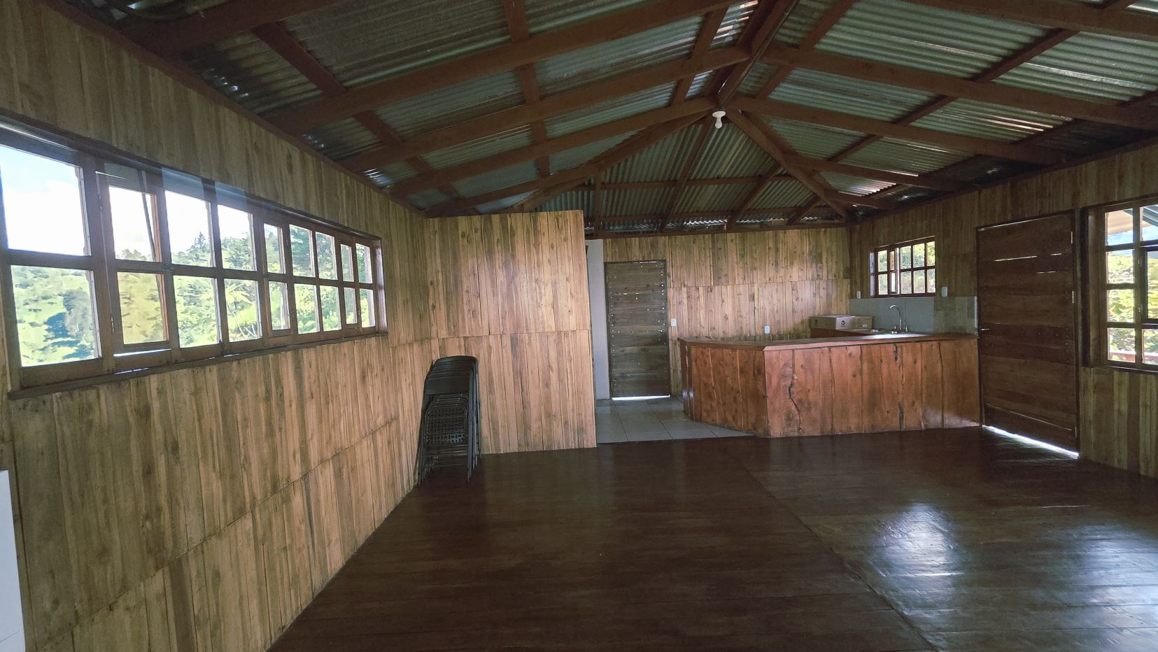 Rustic cabin interior with wooden walls, dark floor, and a central wood-toned kitchen island; large windows reveal greenery outside.