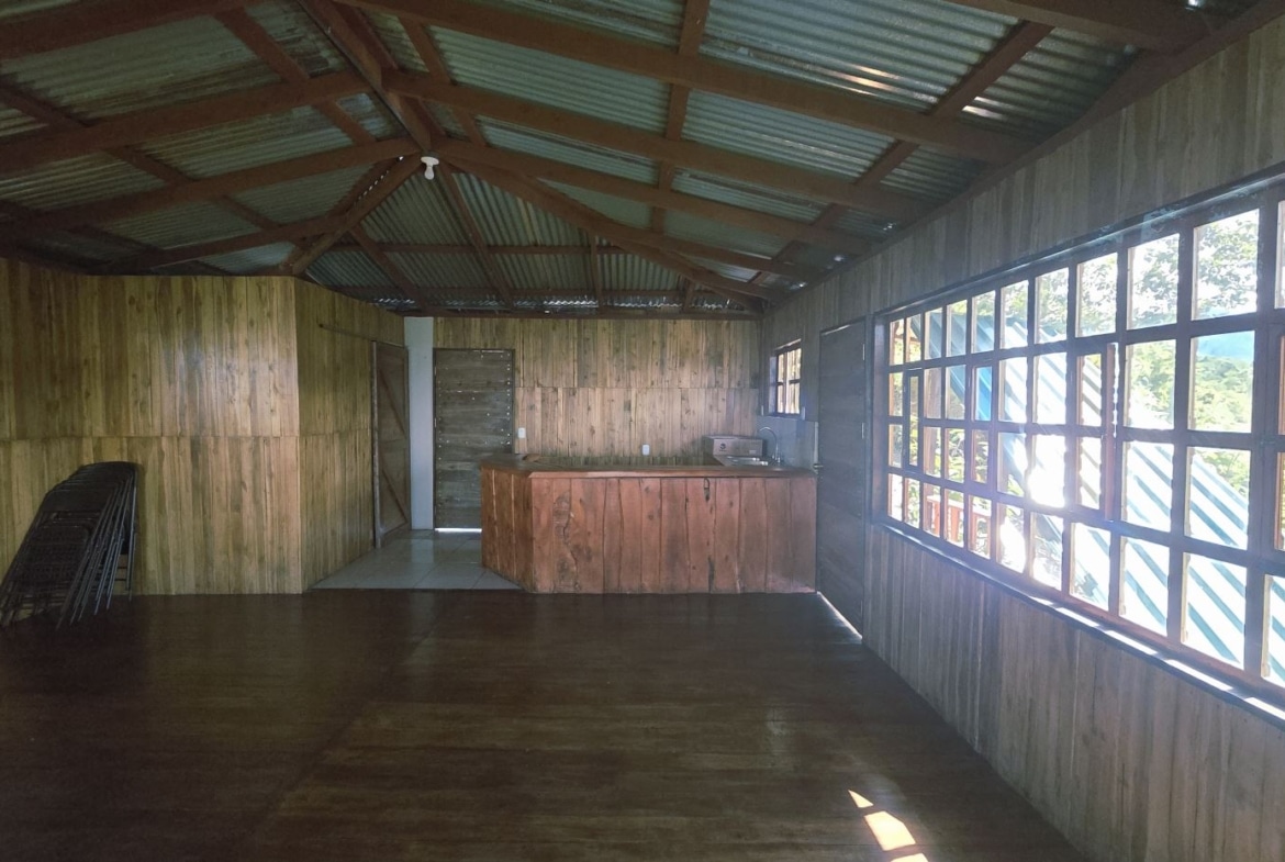 Rustic wooden indoor space with a long wooden counter, large pane windows on the right, and folding chairs stacked in the corner.