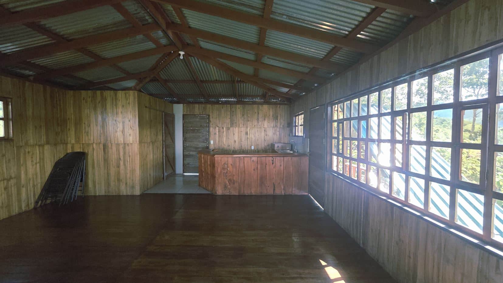 Rustic wooden indoor space with a long wooden counter, large pane windows on the right, and folding chairs stacked in the corner.