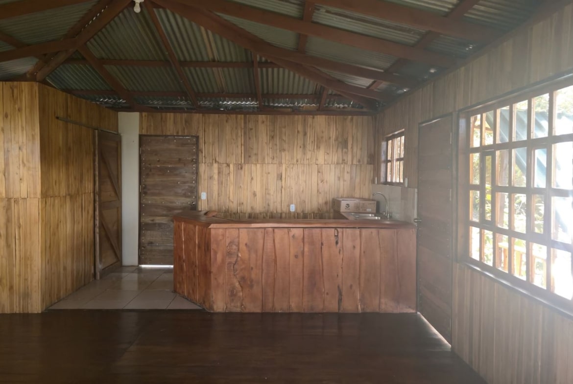 Rustic cabin kitchen with a wooden island, sink, and paneled walls under a metal roof. Large windows run along the right wall.