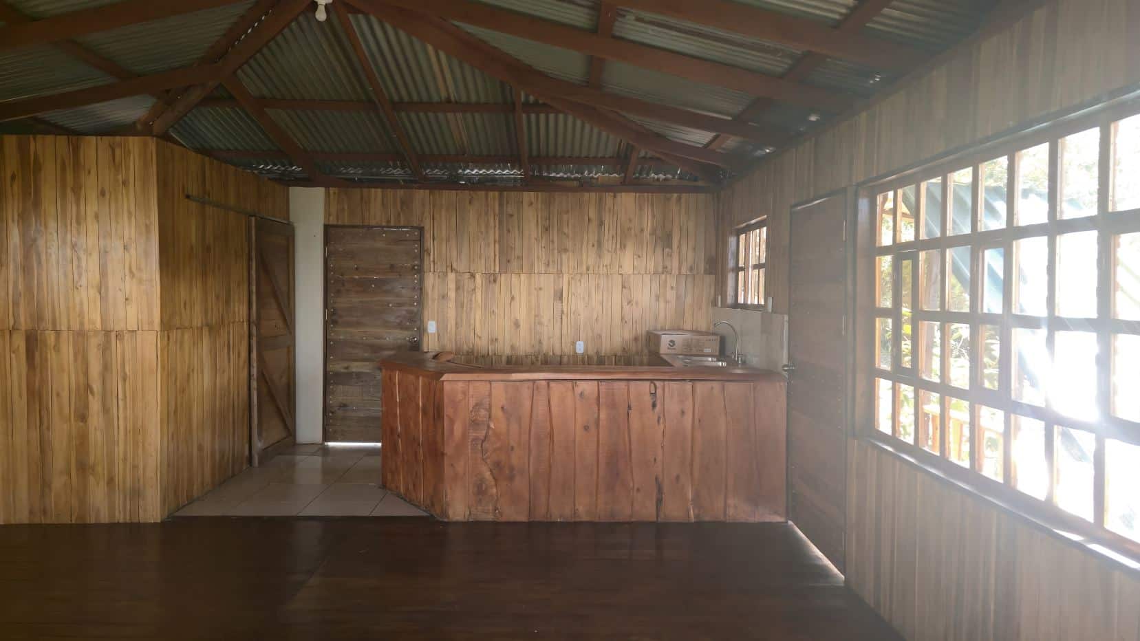 Rustic cabin kitchen with a wooden island, sink, and paneled walls under a metal roof. Large windows run along the right wall.