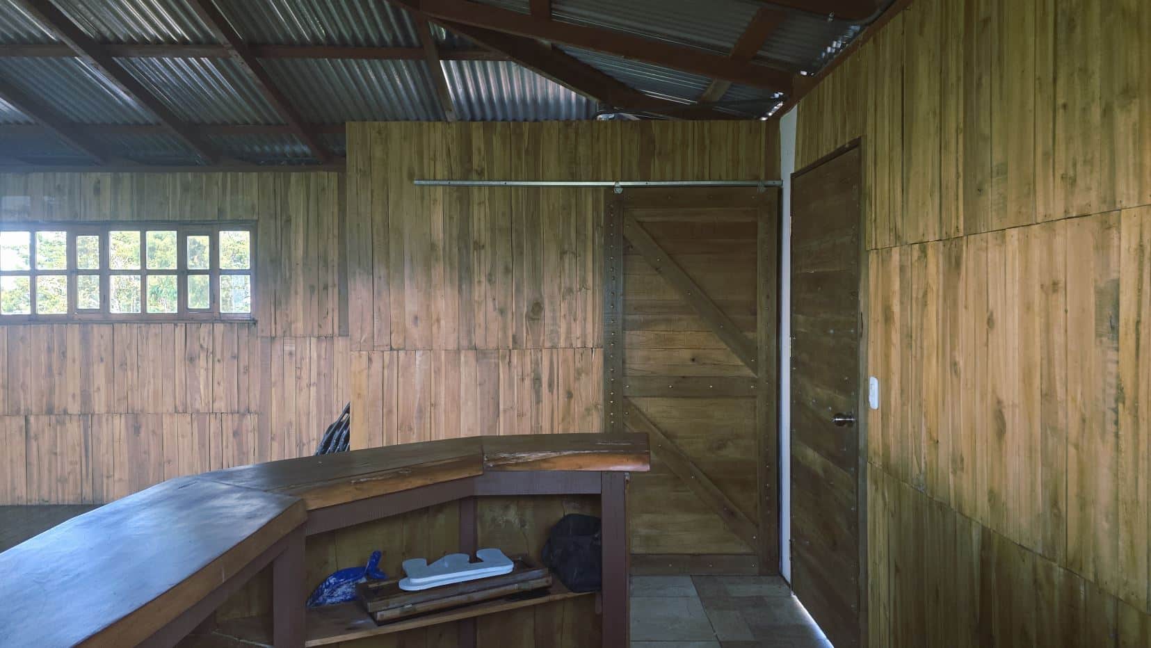 Rustic wooden interior with a curved bar counter in the foreground, wood-paneled walls, a multi-panel window to the left, and a large wooden door on the right side.