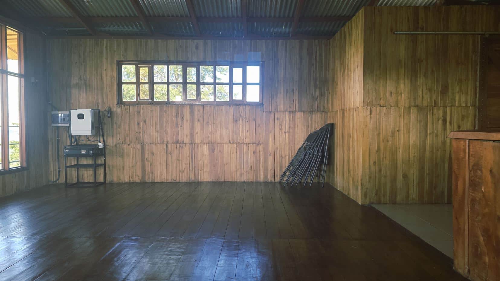 Empty wooden-paneled room with a row of small windows, and a few folding chairs stacked against the right wall near a table with equipment left of center