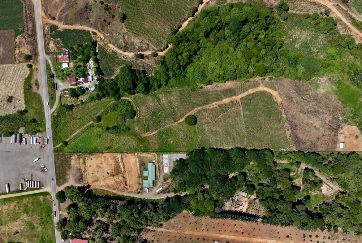 Aerial view of a rural area with fields, roads, and clusters of trees surrounding a few buildings and industrial yards near a main road.