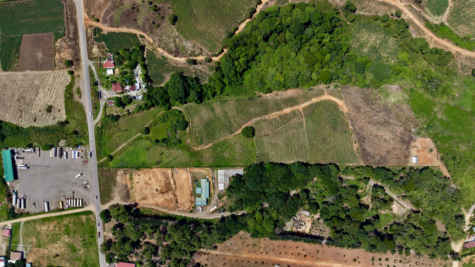 Aerial view of a rural area with fields, roads, and clusters of trees surrounding a few buildings and industrial yards near a main road.