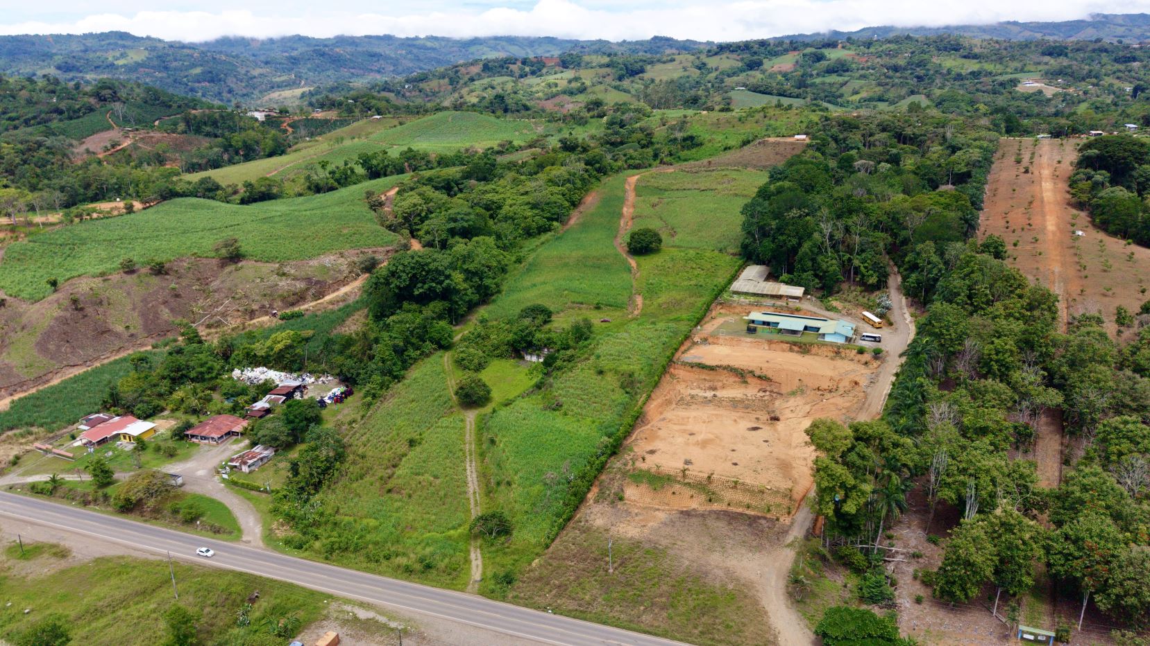 Aerial view of a rural countryside with green fields, small farmhouses along a road, and rolling hills in the distance.