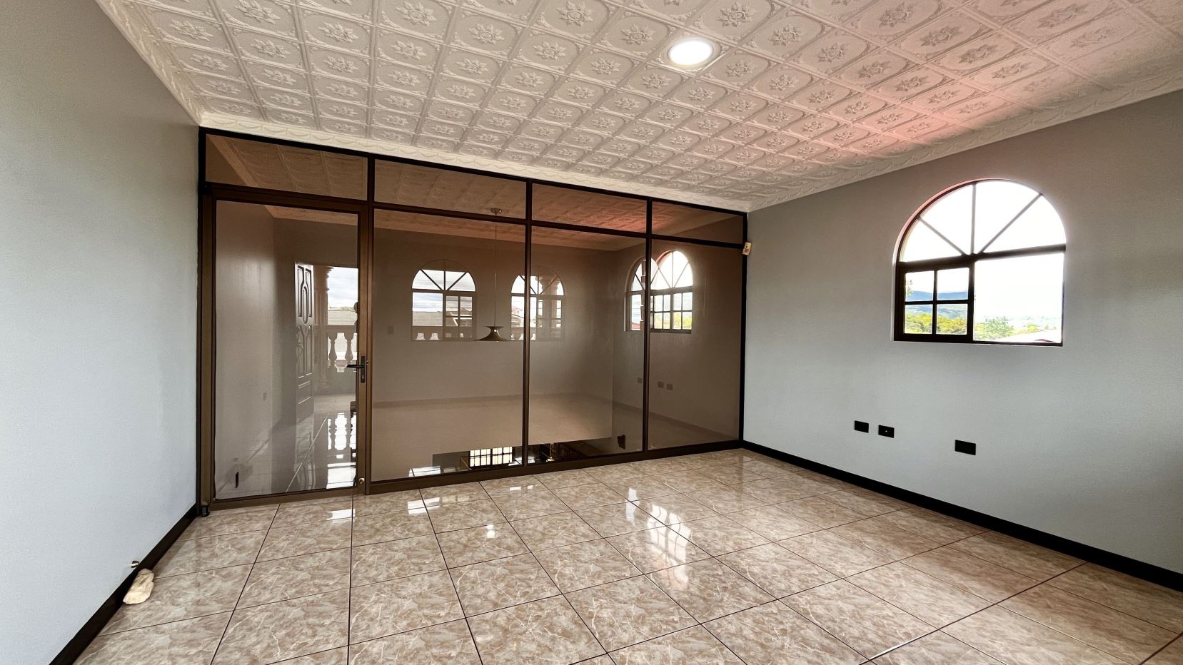 Empty office or living room with marble-tile floor, glass-framed partition and arched window, neutral walls, and decorative patterned ceiling paneling