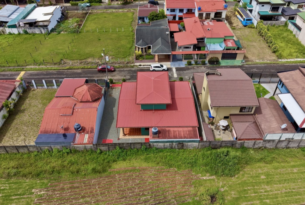 Aerial view of a residential street with red-roofed houses, backyards, and a grassy lot behind, set against a green hillside in the foreground.