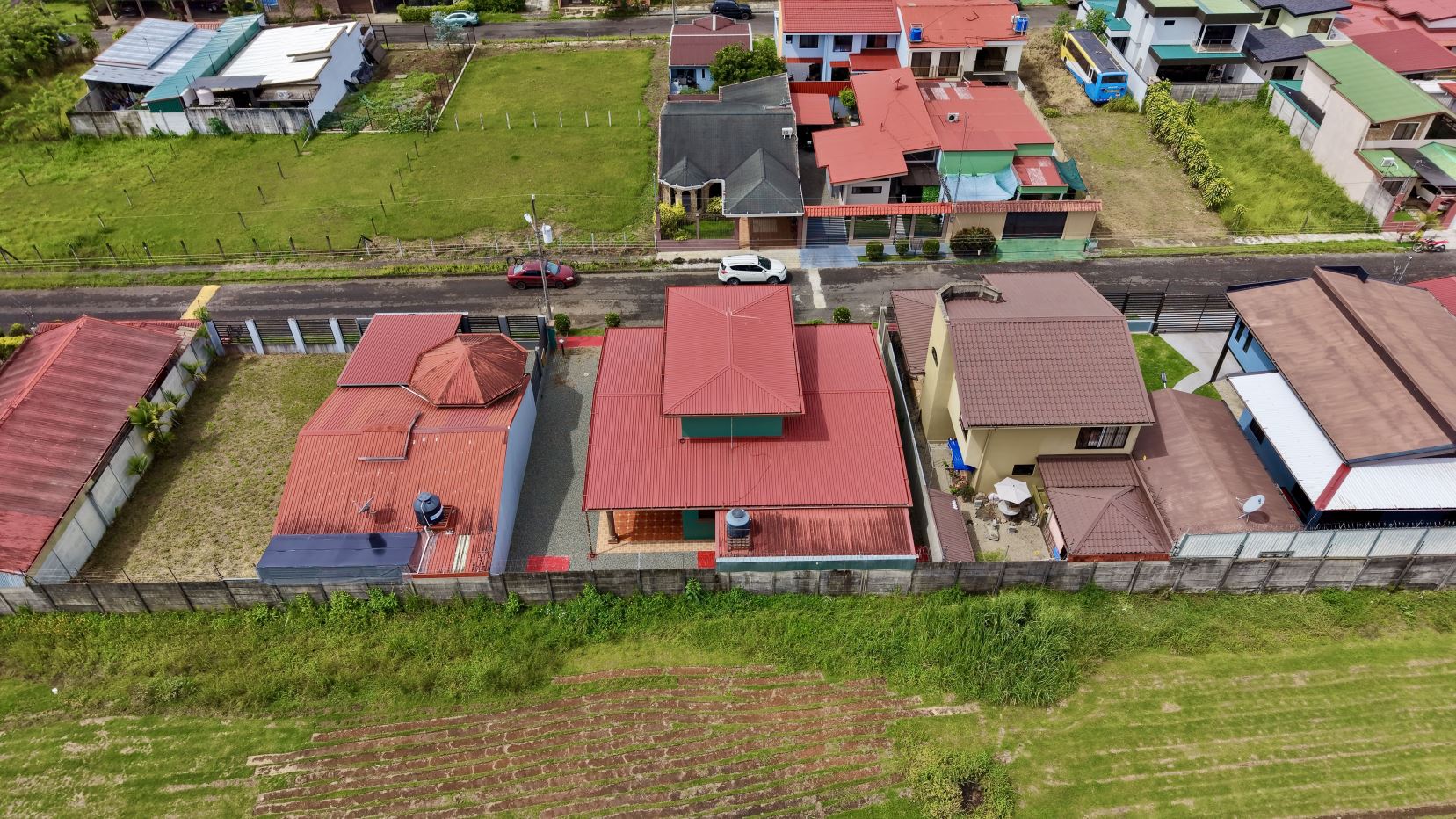 Aerial view of a residential street with red-roofed houses, backyards, and a grassy lot behind, set against a green hillside in the foreground.