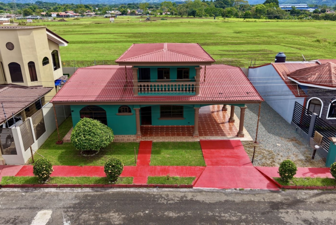 Two-story turquoise house with a red metal roof, balcony, and front porch, set among neighboring homes with green fields beyond.