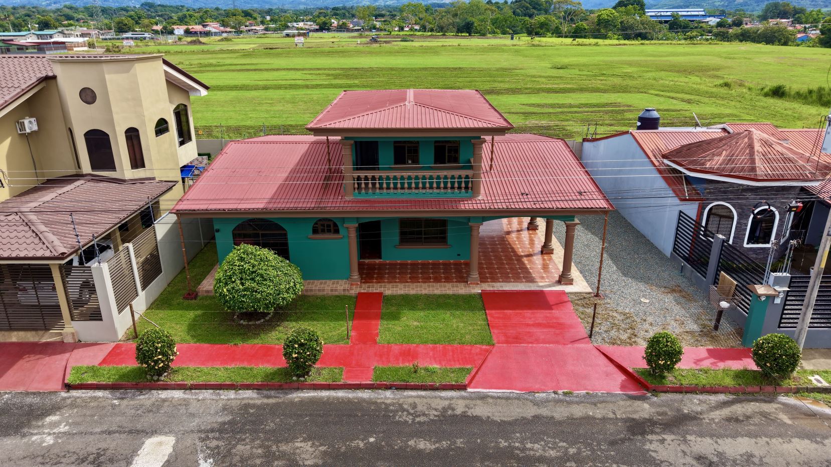 Two-story turquoise house with a red metal roof, balcony, and front porch, set among neighboring homes with green fields beyond.