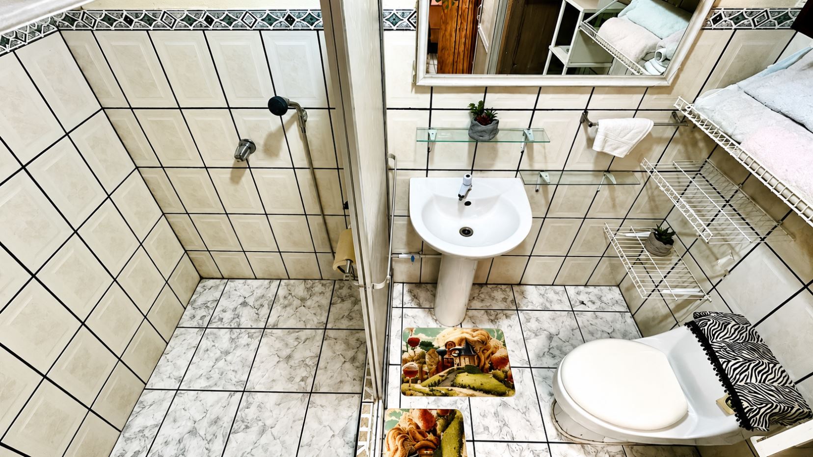 Top-down view of a compact bathroom with a pedestal sink, mirror, toilet, and white wire shelves.