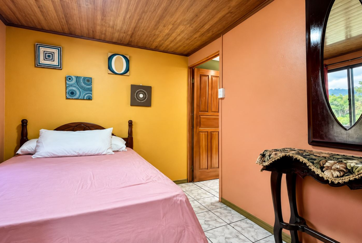 Bedroom with a pink bedspread and white pillows, yellow accent wall with geometric art, wooden ceiling, and doorway to another room.