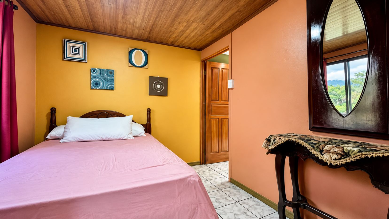Bedroom with a pink bedspread and white pillows, yellow accent wall with geometric art, wooden ceiling, and doorway to another room.