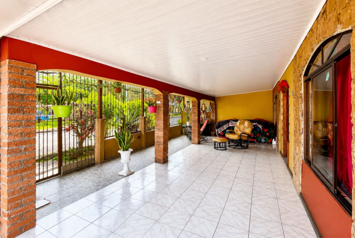 Covered veranda with brick columns, decorative iron railing, and potted plants along a tiled floor. A seating area with chairs is on the right under a white ceiling.