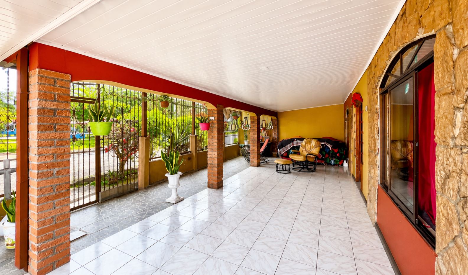 Covered veranda with brick columns, decorative iron railing, and potted plants along a tiled floor. A seating area with chairs is on the right under a white ceiling.