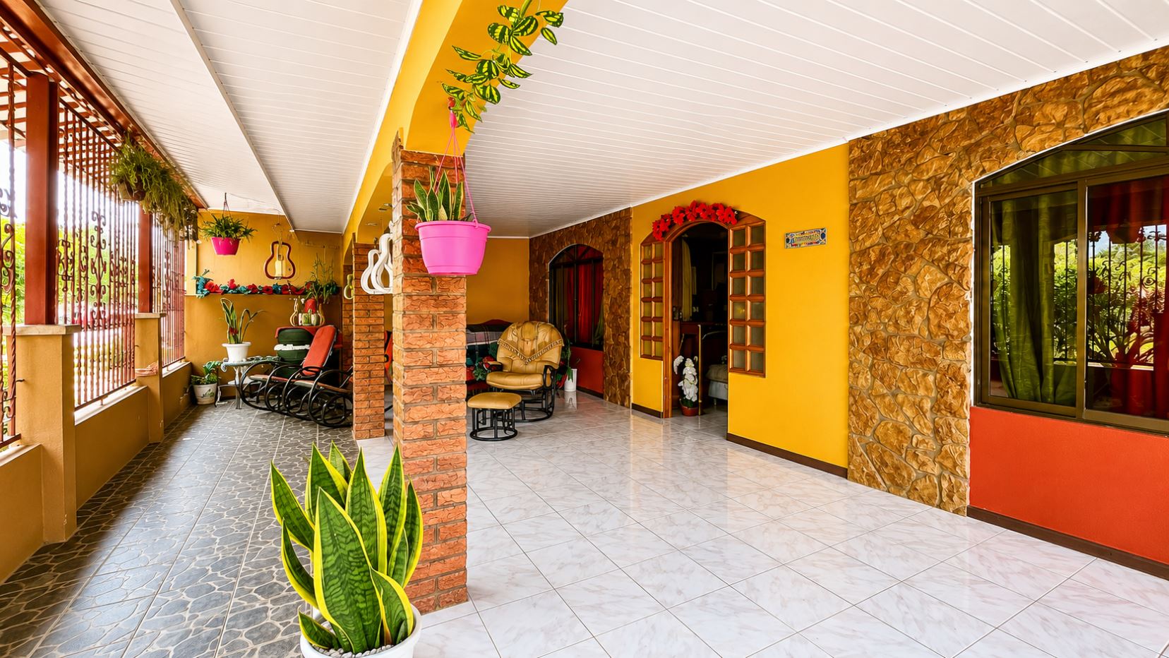 Bright covered patio with yellow walls, stone accents, and tiled floor; potted plants, hanging baskets, and outdoor seating.