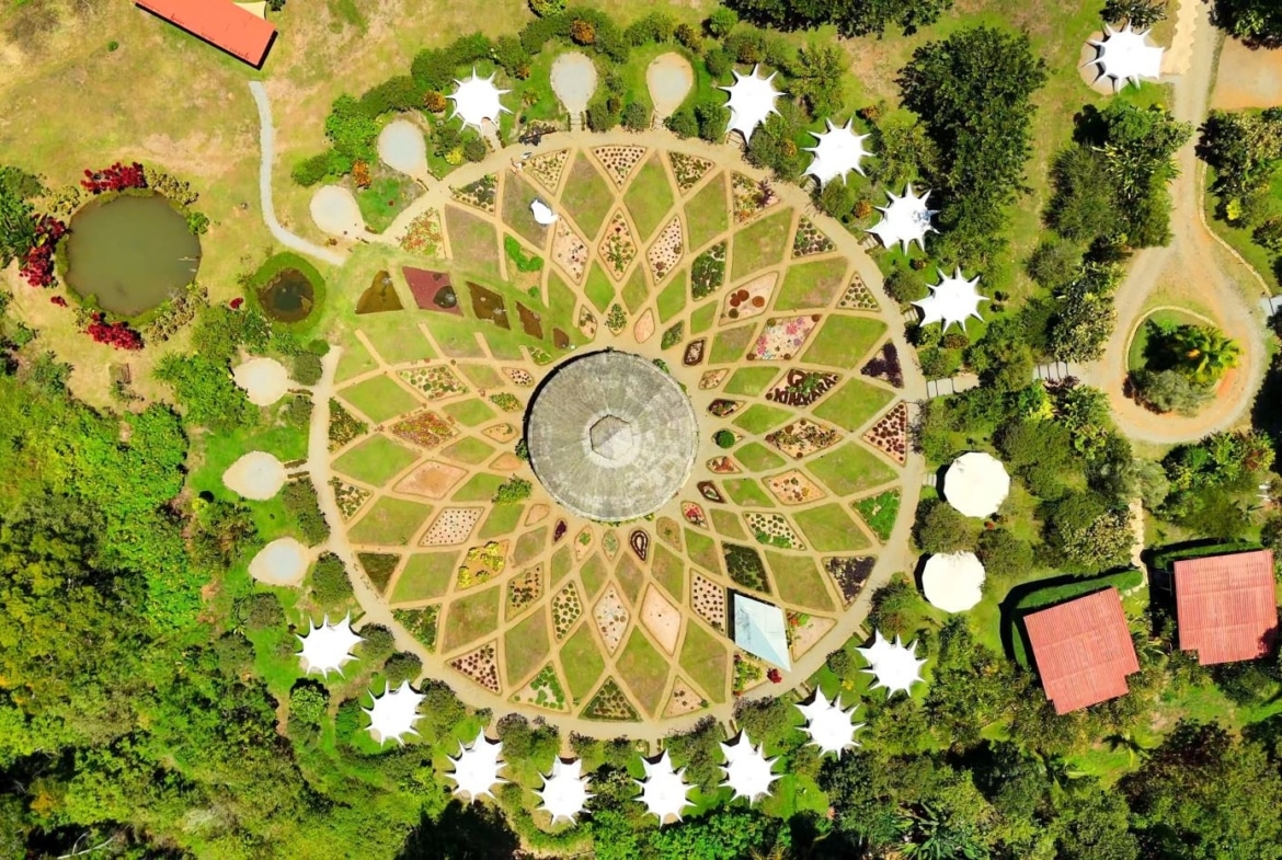 Aerial view of a circular garden with a central domed structure and radial planting beds surrounded by trees and white tents around the edge.