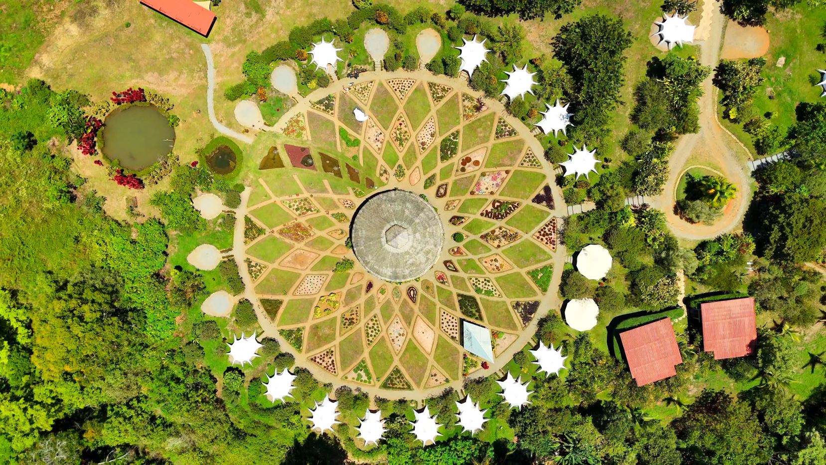 Aerial view of a circular garden with a central domed structure and radial planting beds surrounded by trees and white tents around the edge.
