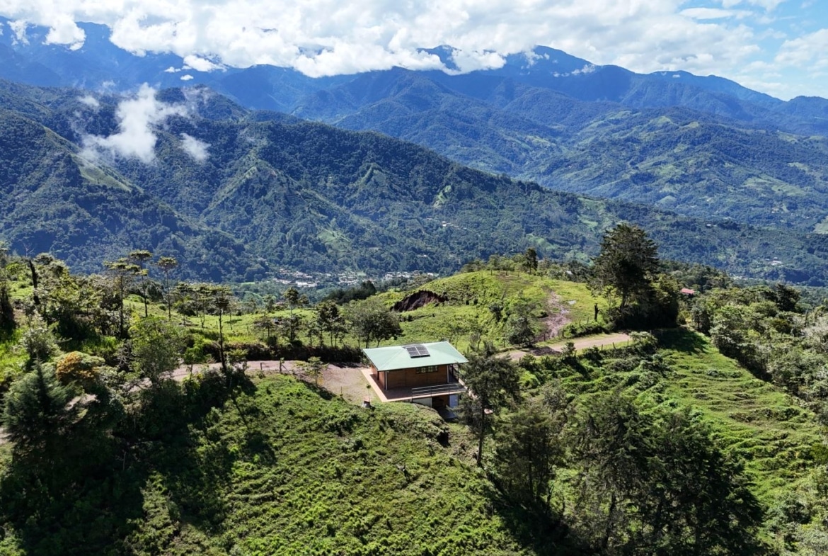 Small cabin with a green roof and solar panels sits on a grassy hillside, with vast green mountains in the distance.