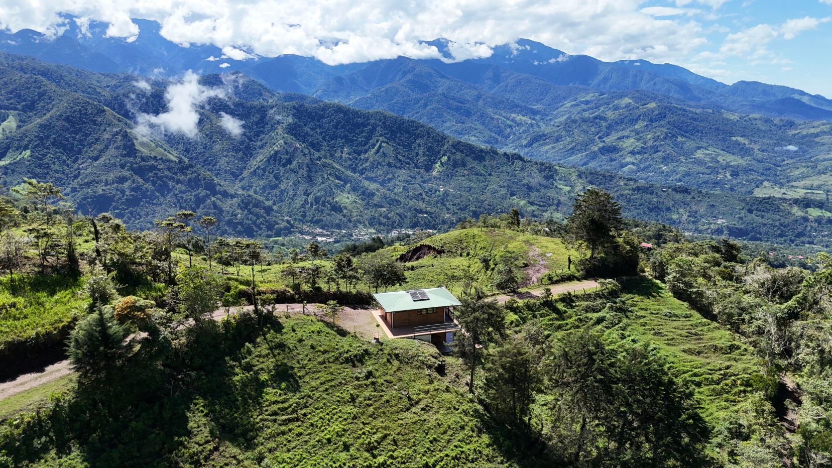 Small cabin with a green roof and solar panels sits on a grassy hillside, with vast green mountains in the distance.
