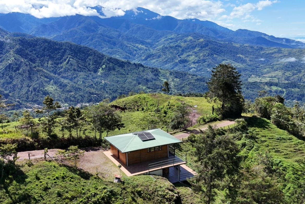 Wooden cabin with a solar panel on its green roof sits on a grassy hillside, with towering blue mountains in the distance.