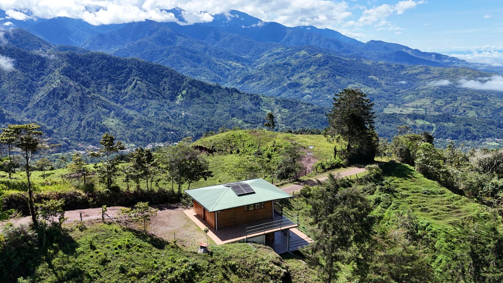Wooden cabin with a solar panel on its green roof sits on a grassy hillside, with towering blue mountains in the distance.