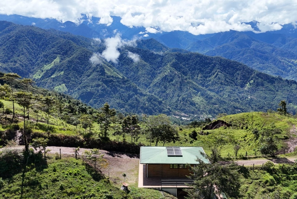 Small cabin with solar panels on a green hillside, overlooking a valley of forested mountains under a blue sky with clouds.