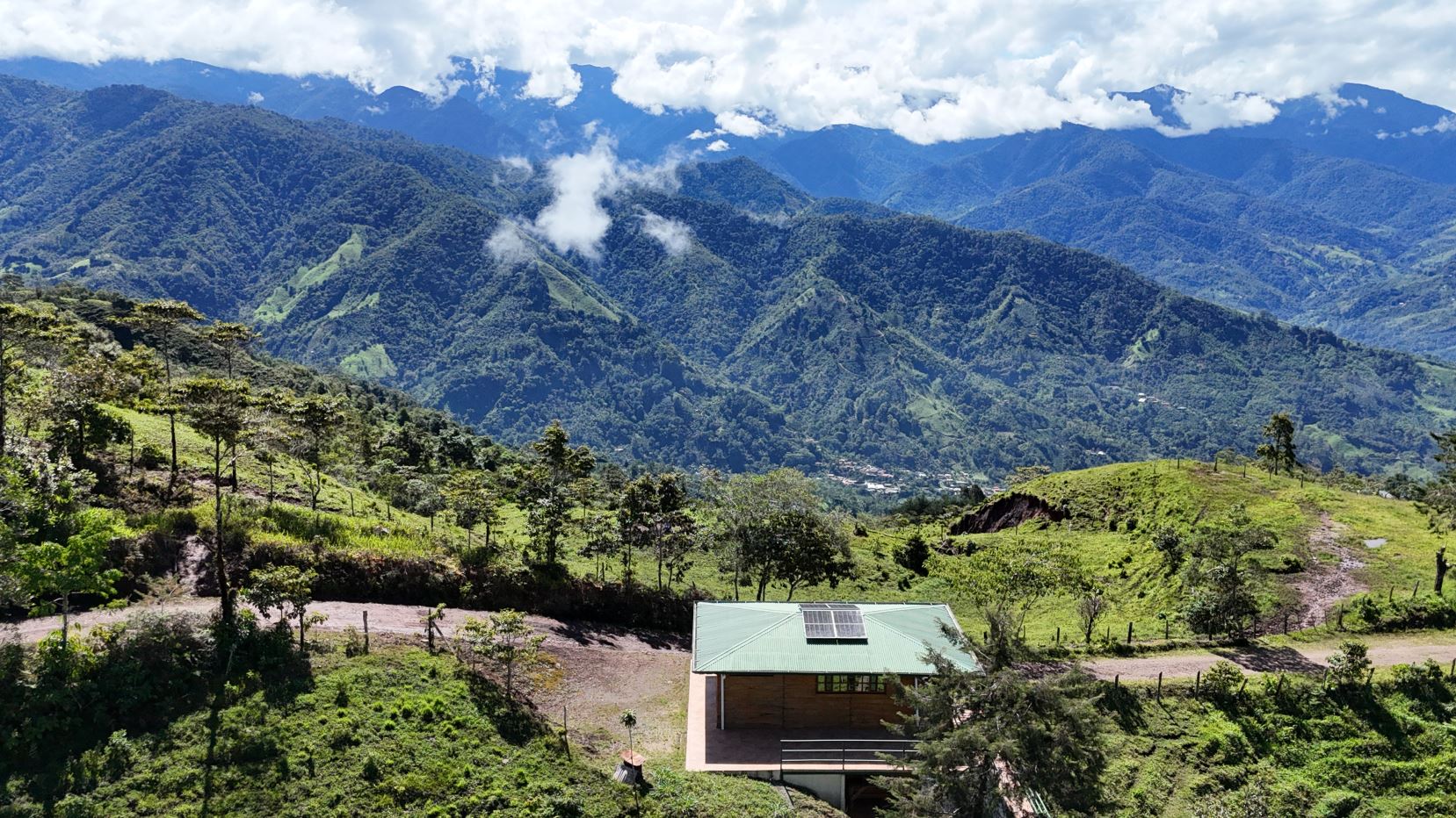 Small cabin with solar panels on a green hillside, overlooking a valley of forested mountains under a blue sky with clouds.