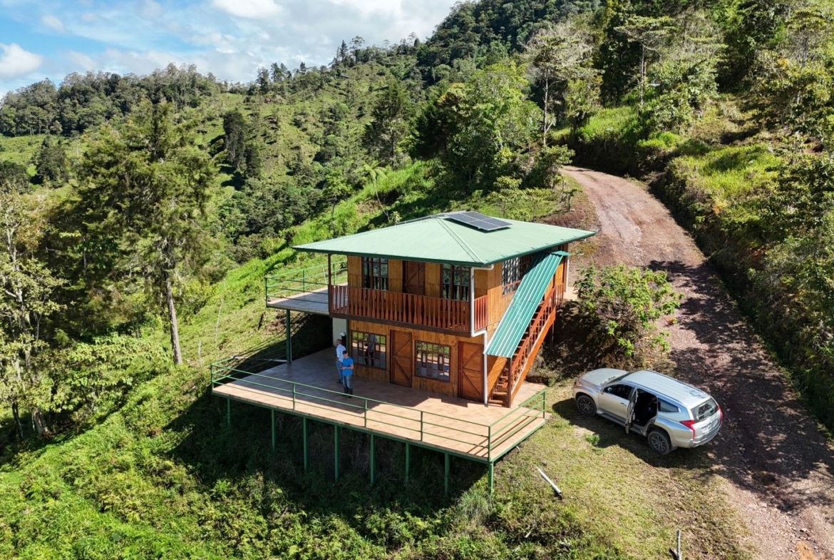 Wooden two-story cabin with green roof on a hillside, solar panel on top, and wraparound deck overlooking a dirt road and forested hills. A silver SUV is parked nearby with a person standing on the deck.
