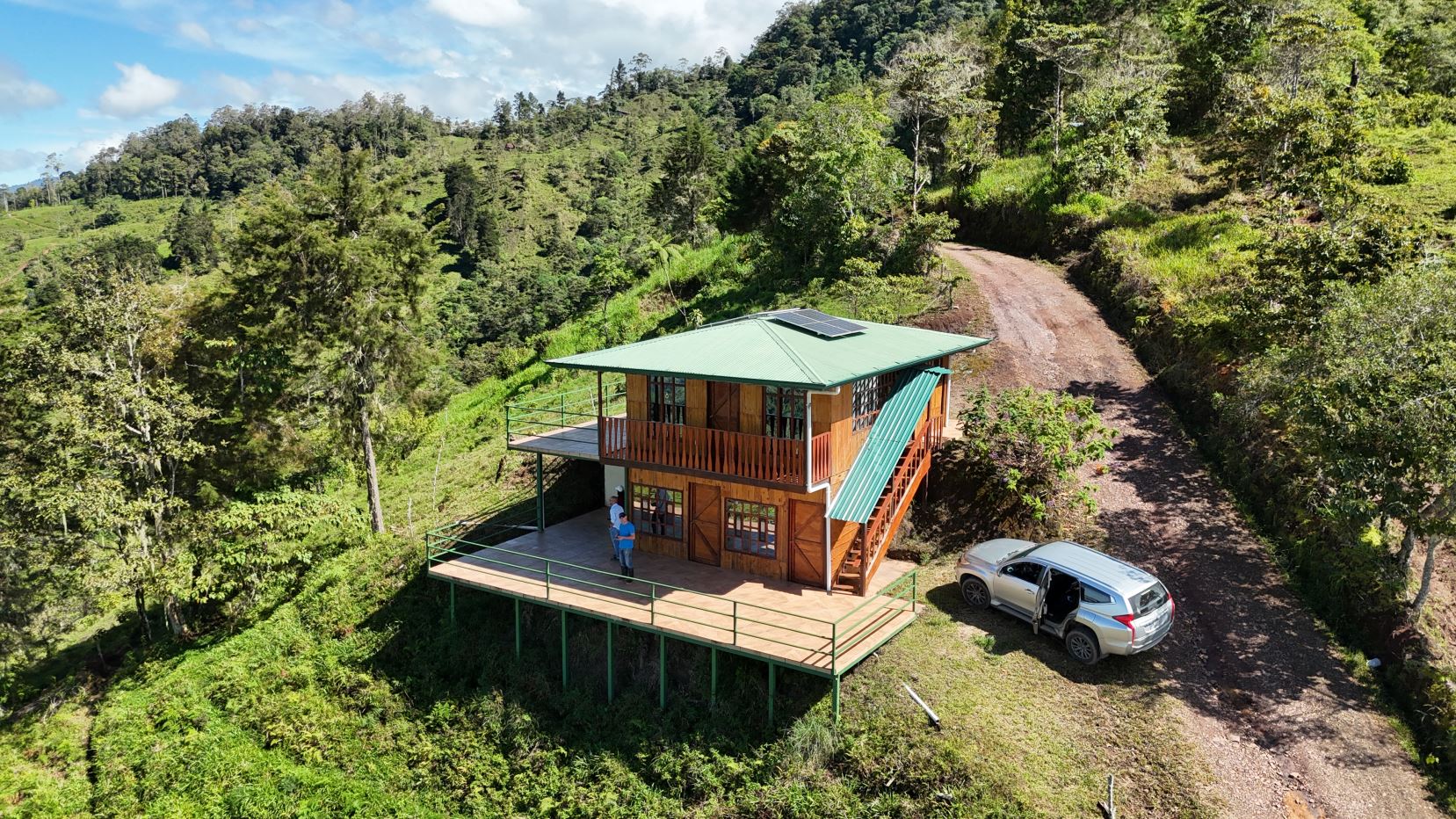 Wooden two-story cabin with green roof on a hillside, solar panel on top, and wraparound deck overlooking a dirt road and forested hills. A silver SUV is parked nearby with a person standing on the deck.