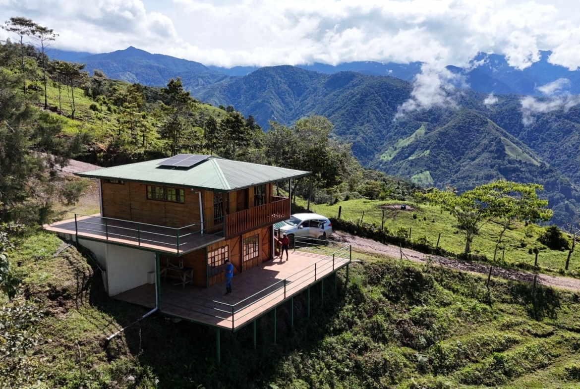 Wooden hillside cabin with a wraparound deck, solar panel on the green roof, and a car parked nearby against a backdrop of lush, misty mountains.